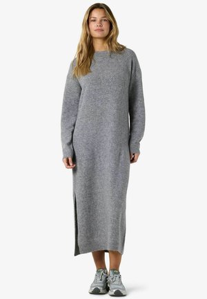 Femme portant une longue robe-pull grise en maille avec des fentes latérales et des chaussures de sport grises, debout devant un fond blanc uni.