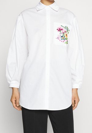 Weißes langärmeliges Hemd mit Knöpfen und einer floral bestickten Tasche, getragen mit schwarzen Hosen vor einem schlichten Hintergrund.
