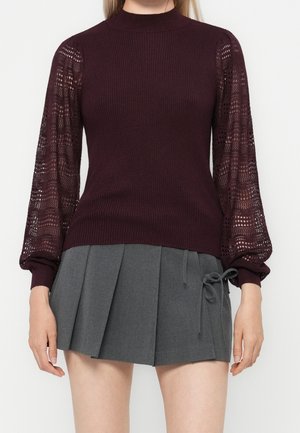 Kvinde iført en bordeaux ribstrikket sweater med blondeballonærmer og en grå plisseret mininederdel med sløjfedetalje i siden.