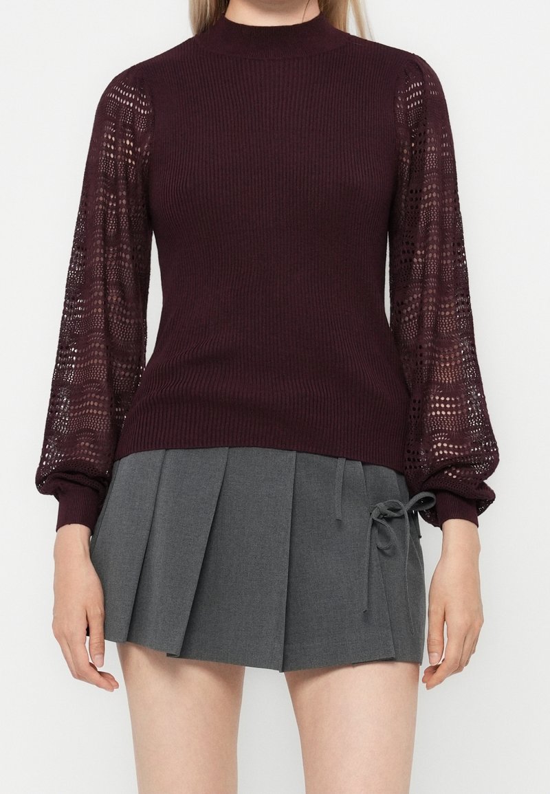 Femme portant un pull côtelé bordeaux avec des manches ballon en dentelle et une mini-jupe plissée grise avec un détail de nœud sur le côté.