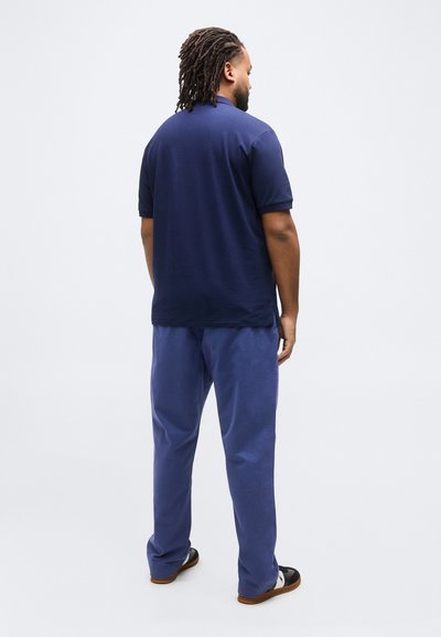 Άνδρας με ντραντλοξ στέκεται με την πλάτη γυρισμένη, φοράει μπλε πόλο πουκάμισο, μπλε παντελόνι και μαύρα-λευκά αθλητικά παπούτσια σε απλό φόντο.