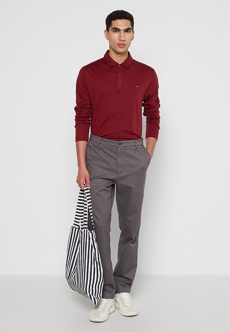 Burgunderfarbener Langarm-Polo mit drei Knöpfen, kombiniert mit grauen Hosen. Das Model hält eine gestreifte Tragetasche und trägt weiße Sneaker.