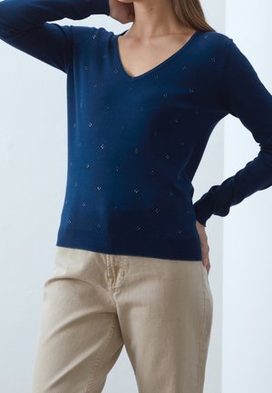 Femme portant un pull en V bleu marine orné de petits clous argentés, associé à un pantalon beige, posant avec une main derrière la tête et l'autre sur la hanche.