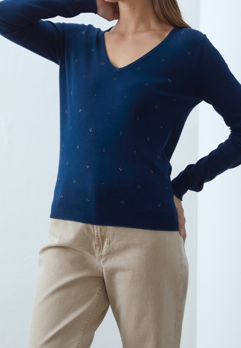 Femme portant un pull en V bleu marine orné de petits clous argentés, associé à un pantalon beige, posant avec une main derrière la tête et l'autre sur la hanche.