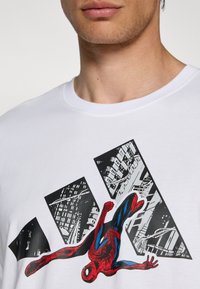 Balta medvilninė marškinėlių su Spider-Man grafika raudona ir mėlyna spalvomis, priešais grafiškus juodos ir baltos miesto peizažų spaudinius. Apvalus iškirpimas.