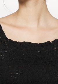 Svart, texturerad topp med fyrkantig halsringning och samlad volangdetalj runt kragen. Materialet visar ett konsekvent, öppet vävmönster.