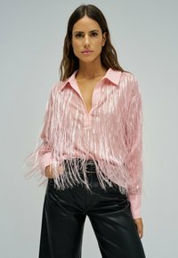 Femme portant un chemisier rose orné de plumes avec un décolleté en V profond et un pantalon en cuir noir, posant devant un fond gris clair uni.