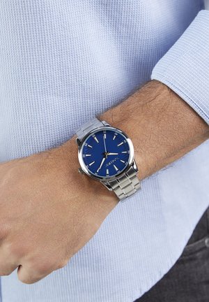 Silberne Metalluhr mit blauem Zifferblatt, versehen mit silberfarbenen Stundenmarkierungen und Zeigern, getragen an einem Handgelenk mit einem strukturierten blauen Hemd.