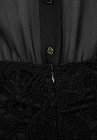 Abito nero con parte superiore trasparente, abbottonato con accenti dorati a forma di stella, e una parte inferiore in pizzo testurizzato con dettagli in zip.