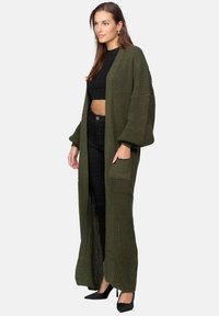 Lång, öppen cardigan i olivgrön stickning, med breda ärmar, ribbat mönster och en framlappad ficka. Matchad med svarta högmidjade byxor.