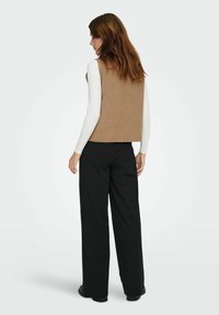 Donna con capelli castani che indossa un gilet beige senza maniche sopra una maglia bianca a maniche lunghe e pantaloni neri a gamba larga, in piedi di spalle.