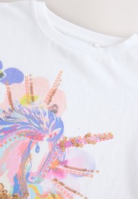 Chemise blanche avec imprimé licorne coloré ornée de paillettes dorées brillantes et d'accents floraux pastel près du col.
