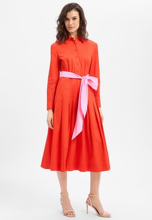 Femme portant une robe midi orange à manches longues et boutonnée, avec une ceinture rose nouée à la taille et des sandales à talons hauts couleur nude.