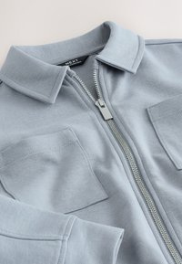 Veste bleu clair à fermeture éclair avec col et deux poches poitrine, avec une fermeture éclair argentée et une étiquette visible à l'intérieur du col.