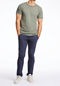 Mężczyzna ubrany w oliwkowozielony t-shirt z krótkim rękawem, granatowe spodnie i białe trampki, stoi z rękami w kieszeniach na białym tle.