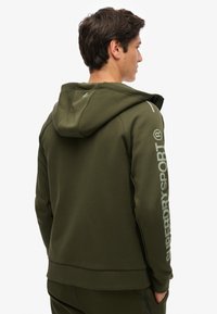 Felpa verde oliva con cappuccio, caratterizzata da un grande logo lungo la manica sinistra, dettagli cuciti sul retro e una texture liscia.