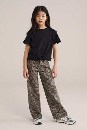 Młoda dziewczyna stoi, ubrana w czarną bluzkę z krótkim rękawem związaną w talii, szerokie spodnie w panterkę oraz białe trampki na beżowym tle.
