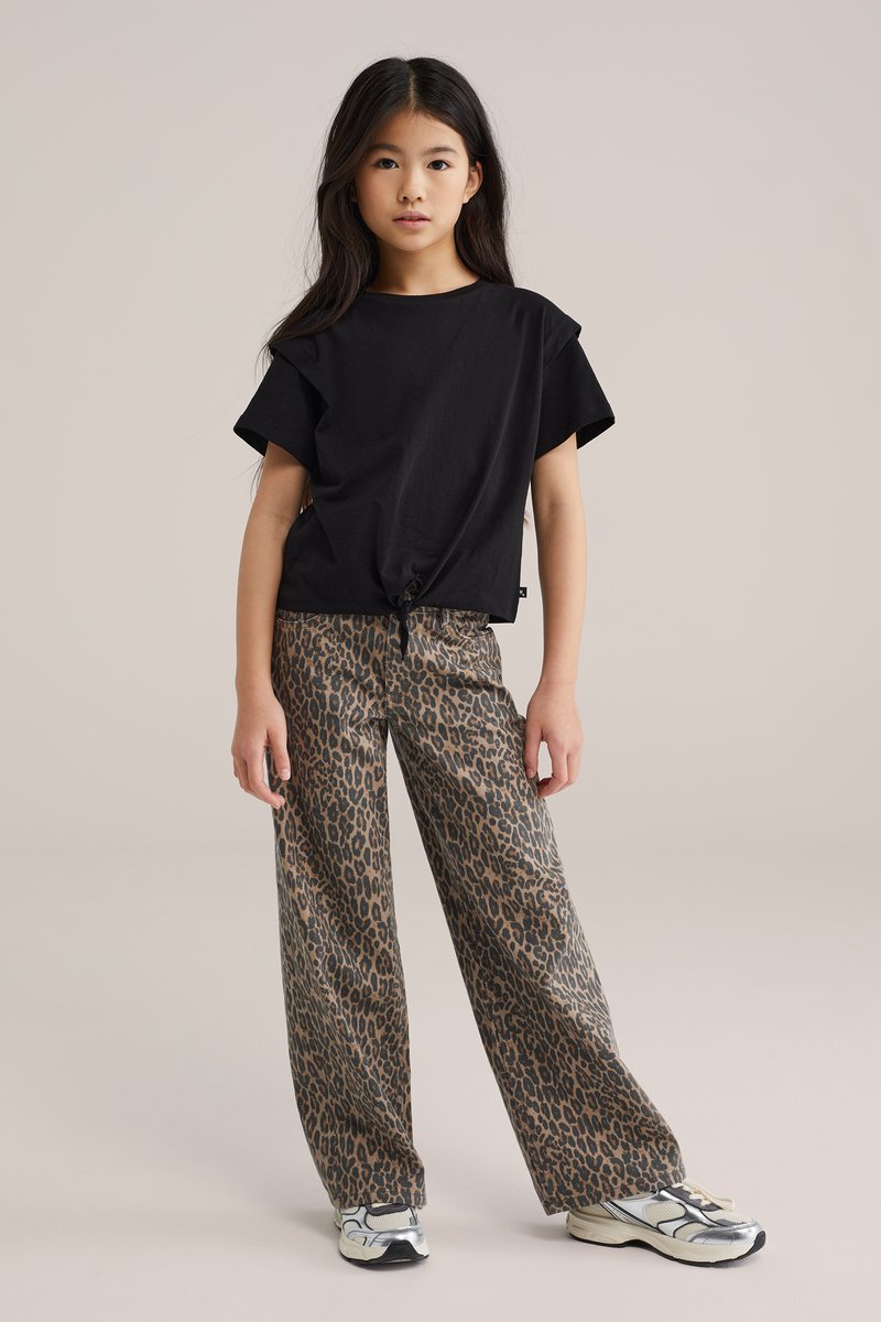 Junges Mädchen steht und trägt ein schwarzes kurzärmeliges Oberteil, das an der Taille gebunden ist, weit geschnittene Hose mit Leopardenmuster und weiße Sneaker vor beigem Hintergrund.