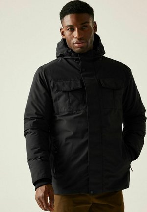 Mand med mørk hud iført en sort hættejakke til vinter og brune bukser, stående mod en neutral baggrund.