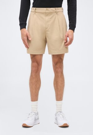 Mand iført beige plisserede shorts, hvide sokker, hvide sneakers og en sort langærmet skjorte, står foran en hvid baggrund.