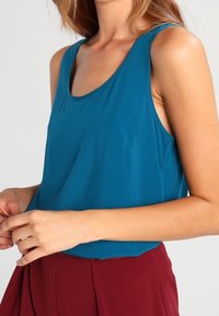 Haut sans manches bleu sarcelle en tissu doux, avec un col rond et une coupe décontractée, associé à un pantalon taille haute bordeaux.