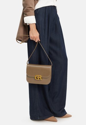 Kobieta trzymająca małą torebkę z taupe skóry z złotym logo, ubrana w szerokie ciemnoniebieskie spodnie, beżowe szpiczaste buty i brązową kurtkę.