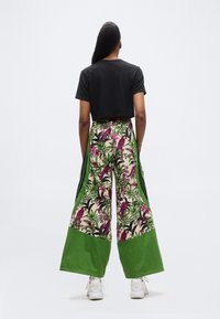 Persona con trenzas largas que lleva una camiseta negra corta, pantalones de pierna ancha con estampado verde y de aves tropicales, y zapatillas blancas, vista desde atrás.