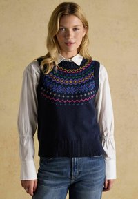 Tumši zils adīts žakets ar krāsainiem ģeometriskiem rakstiem ap apkakli, virs balta krekla ar apkakli, kopā ar ziliem džinsiem.