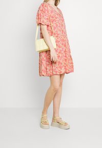 Rosa blomsterklänning med korta puffärmar, tillsammans med en gul vadderad axelväska och beige plattformssandal med gula remmar.