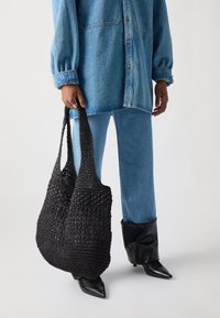 Borsa tote nera intrecciata con due lunghe cinghie, caratterizzata da una superficie testurizzata e fondo arrotondato, abbinata a un outfit in denim e scarpe a punta.