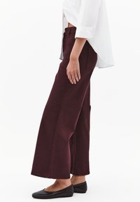 Jeans larghi bordeaux in denim, con vita alta, tasche laterali e linee pulite, abbinati a una camicia bianca con bottoni e ballerine nere.