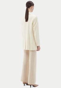 Blazer crème à coupe ajustée, associé à un pantalon large en maille de couleur beige clair texturée. La tenue est complétée par des escarpins pointus.