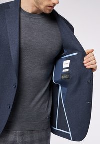 Blazer blu Navy con tessuto testurizzato, fodera azzurro chiaro e etichetta interna. Indossato sopra un maglione grigio, che mette in risalto una vestibilità su misura.