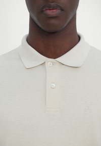 Gros plan d'une personne portant un polo beige clair avec un col et deux boutons fermés.