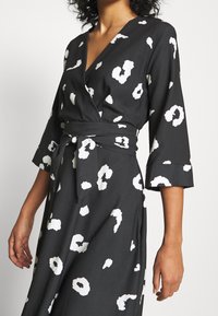 Kvinna i en svart omslagsklänning med vita abstrakta prickar, trekvartsärmar och ett knytbälte i midjan.