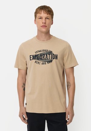 Ung mand med kort lyst brunt hår iført en beige t-shirt med teksten "Established for Exploration Here and Now" og sorte bukser.