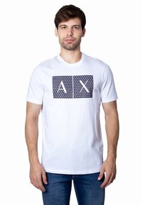 Valge puuvillane T-särk, millel on ees geomeetriline mereväe sinine graafika ja "AX" tähed. Lühikesed varrukad ja ümmargune kaelus.