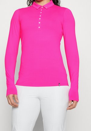 Persoon draagt een felroze poloshirt met lange mouwen, witte knopen en een witte broek tegen een effen achtergrond.