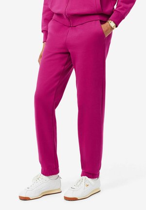 Personne portant un pantalon de jogging rose vif, des baskets blanches avec semelles en gomme, et une veste zippée rose assortie, main dans la poche.