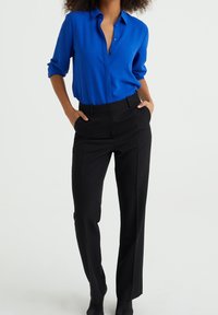 Blå knäppskjorta med uppvikta ärmar, kombinerad med svarta skräddarsydda byxor. Skjortan har en slät textur; byxorna har rak benvidd.