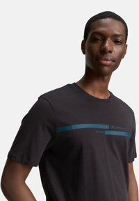 Schwarzes Baumwoll-T-Shirt mit kurzen Ärmeln, das einen horizontalen türkisen Streifen und den auf der Brust aufgedruckten Text "EST. 2007" in Blau zeigt.