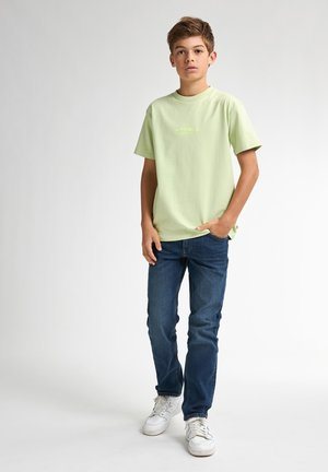 Lichtgroene katoenen t-shirt met korte mouwen en een klein logo, gecombineerd met blauwe denim jeans en witte sneakers. Eenvoudige, casual outfit.