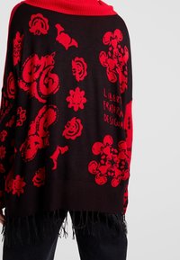 Zwarte gebreide poncho met rode bloemen- en abstracte patronen, voorzien van een rode geribde kraag en een zwarte zoom met franjes.