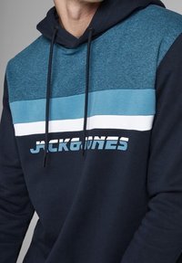 Laivastonsininen huppari, jossa on teksturoitu yläosa teal-värissä, valkoisia ja vaaleansinisiä vaakaraitoja sekä rohkeilla kirjaimilla varustettu logo.