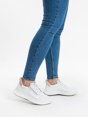 Person trägt blaue enge Jeans und weiße Turnschuhe mit seitlichem Reißverschluss, steht auf einer weißen Oberfläche.