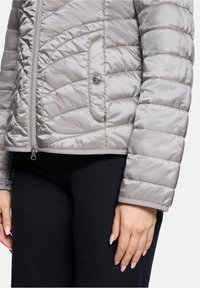 Veste matelassée gris clair en matériau brillant, avec une fermeture éclair, une poche latérale à bouton et des poignets de manches côtelés.