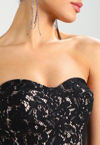 Mustaton pitsiverhoiltu mekko, jossa on kukkakuviot, istuva korsetti ja monimutkaiset yksityiskohdat. Pitkät kimaltavat roikkuvat korvikset korostavat ilmettä.