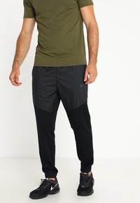 Mustat ja harmaat urheiluvaatteet, joissa on kapeneva muotoilu, sileä pinta ja sivutaskut. Yhdistetty tiukkaan oliivinvihreään t-paitaan.