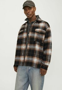 Black and brown plaid flannel jacket with two chest pockets, button closure, and collar detail, layered over a dark shirt and denim jeans.