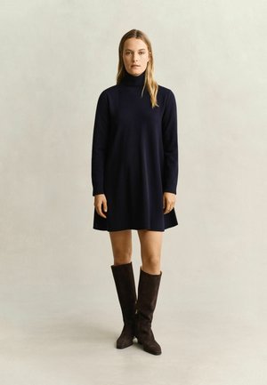 Femme debout portant une robe noire à manches longues et col roulé ainsi que des bottes hautes marron foncé sur un fond neutre.